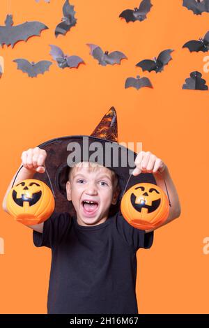 Joyeux Halloween.Halloween effrayante pour les enfants.Garçon dans un chapeau tenant une citrouille d'halloween devant sa tête sur un fond orange avec des battes en papier, verticales Banque D'Images