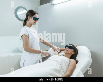 Jeune maître dans les lunettes faisant la procédure d'enlèvement de cheveux laser avec l'équipement professionnel en main dans le salon de lumière Banque D'Images