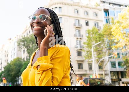 Une femme afro-américaine positive en lunettes qui regarde loin tout en ayant une conversation téléphonique dans la rue avec des bâtiments résidentiels et des arbres dans la ville Banque D'Images
