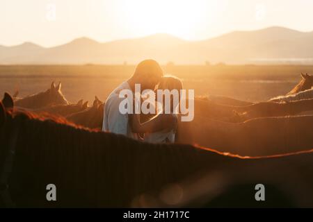 Homme embrassant femme tendre debout à proximité les uns des autres parmi les chevaux calmes dans la campagne vallonnée à la lumière du coucher du soleil Banque D'Images