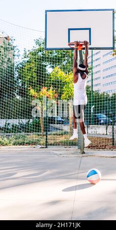 Vue arrière d'un joueur de basket-ball afro-américain anonyme suspendu sur un panier en métal sur le terrain de jeu en été Banque D'Images