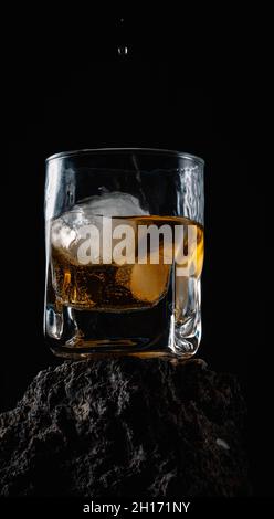 Gouttes de whisky tombant sur des glaçons servis dans du verre cristal placé sur une surface rugueuse sur fond noir Banque D'Images