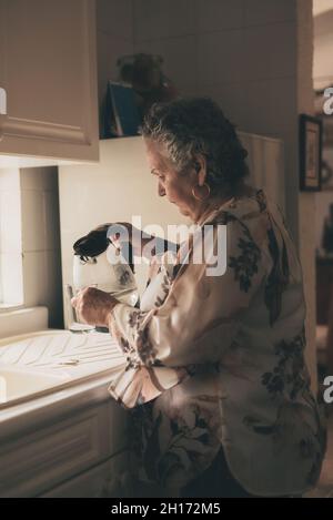 Vue latérale d'une femme âgée concentrée versant de l'eau chaude de la bouilloire électrique transparente dans une tasse de thé en verre tout en faisant des boissons avec thé dans la cuisine Banque D'Images
