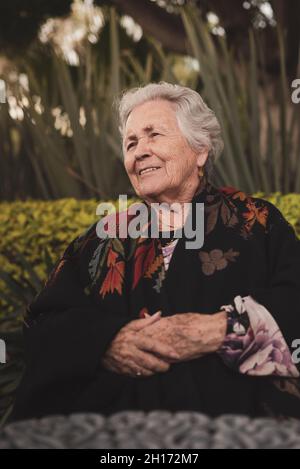Vue latérale d'une femme âgée avec cheveux gris enveloppés dans un tissu écossais assis à une table de jardin et vue sur l'extérieur par temps ensoleillé Banque D'Images