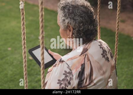 Vue arrière de la dame portant un chemisier assise dans le parc et lisant un livre électronique Banque D'Images