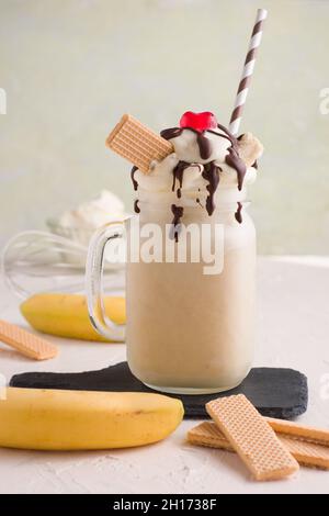Pot en verre de Milk-Shake à la banane sucrée recouvert de gaufres à la crème fouettée, chocolat et cerise sur la planche à découper Banque D'Images
