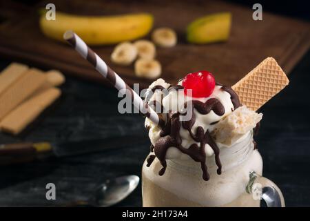 Du dessus du pot en verre de Milk-Shake de banane sucrée recouvert de gaufres à la crème fouettée, chocolat et cerise sur la table Banque D'Images