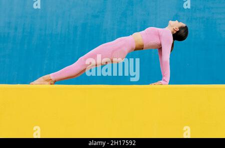 Vue latérale d'une femme mince portant un activewear rose clair pratiquant le yoga asana Purvottanasana debout dans la planche du haut sur fond bleu et jaune Banque D'Images