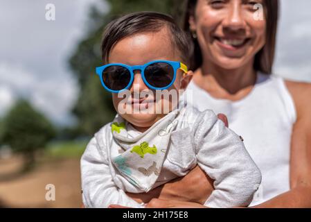 Crop positive mère tenant enfant mignon dans des lunettes de soleil tout en se tenant dans la zone rurale avec de grands arbres verts sur fond flou Banque D'Images