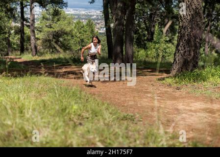 Tout le corps d'une femme active qui court sur la route rurale avec un chien fidèle tout en s'entraîner à grove le jour de l'été Banque D'Images