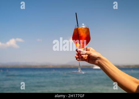 Main féminine tenant un verre de cocktail Aperol Spritz sur fond de mer Banque D'Images