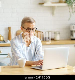 Femme sénior concentrée écrivant quelque chose sur ordinateur portable tout en travaillant en ligne à partir de la maison Banque D'Images