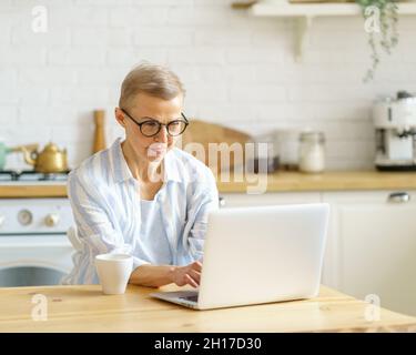 Femme sénior concentrée écrivant quelque chose sur ordinateur portable tout en travaillant en ligne à partir de la maison Banque D'Images