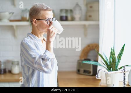 Belle femme mûre commençant le jour avec le café tout en regardant à la fenêtre dans la cuisine Banque D'Images