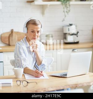 Femme mûre concentrée dans un casque assis dans la cuisine et de prendre des notes, étudiant en ligne sur un ordinateur portable Banque D'Images