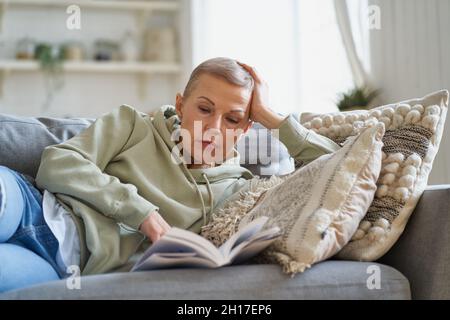 Joyeuse femme heureuse lisant le livre et souriant tout en se relaxant sur le canapé à la maison Banque D'Images