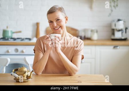 Belle femme mûre commençant le jour avec le café tout en étant assis à une table en bois dans la cuisine Banque D'Images
