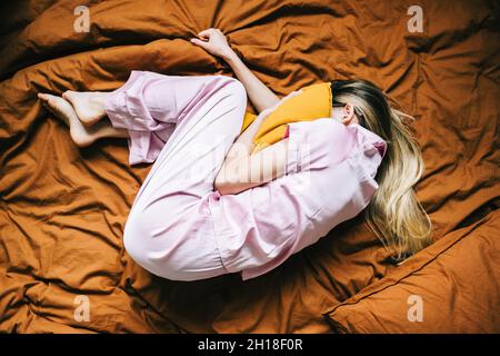 Femme du millénaire du caucase déprimée, couchée seule sur le lit et couvrant la tête avec un oreiller sensation de peur, vue du dessus. Banque D'Images