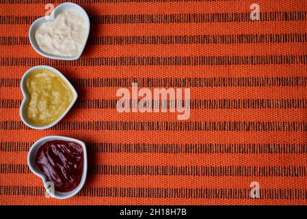 Petits bols en forme de cœur avec ketchup, mayonnaise et sauce moutarde isolés d'un côté sur un fond orange rugueux.espace de copie. Banque D'Images