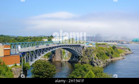 Pont Reversing Falls à Saint John, Nouveau-Brunswick, Canada, vu du parc Wolastoq.Le brouillard glisse sur le centre-ville au loin.Jour ensoleillé. Banque D'Images