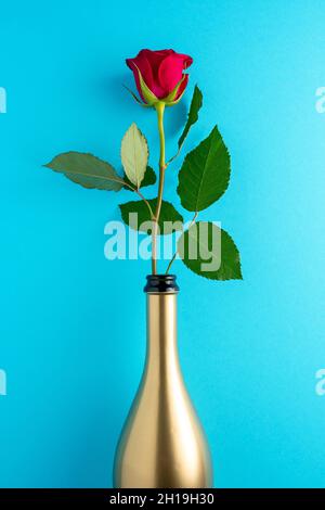 Rose rouge en bouteille de champagne dorée sur fond bleu.Carte de vœux pour la Saint-Valentin, la Fête des femmes.Concept de vacances minimal. Banque D'Images