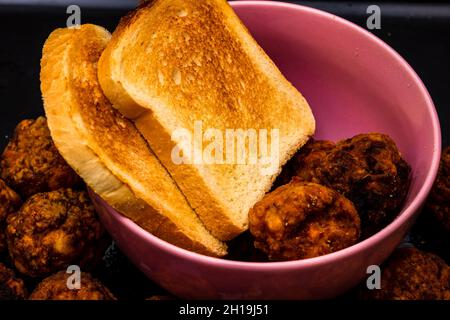 Un gros plan de boulettes de viande fraîchement frites dans un bol rose avec des tranches de pain grillées ; fond noir Banque D'Images