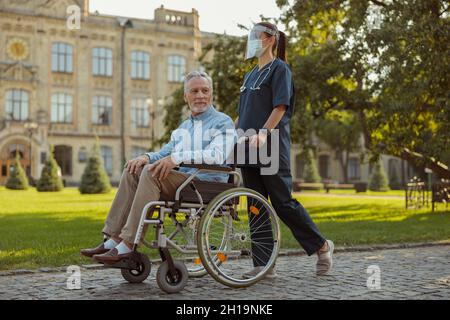 Prise de vue en longueur d'une jeune infirmière soignante portant un écran facial et un masque marchant avec un patient adulte en fauteuil roulant Banque D'Images
