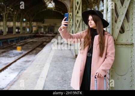 Une jeune femme heureuse sur la plate-forme de la gare en manteau rose et chapeau noir attend le train et communique par communication vidéo avec des parents en ligne ou prend un selfie sur un smartphone Banque D'Images