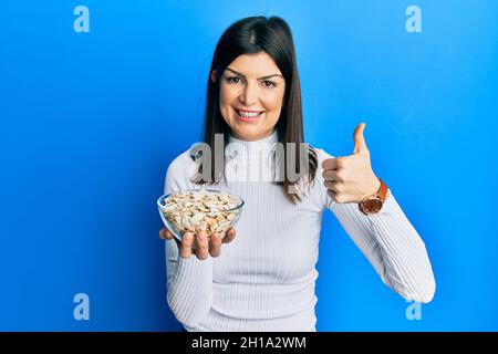 Jeune femme hispanique tenant des graines de citrouille bol souriant heureux et positif, pouce vers le haut faisant excellent et signe d'approbation Banque D'Images