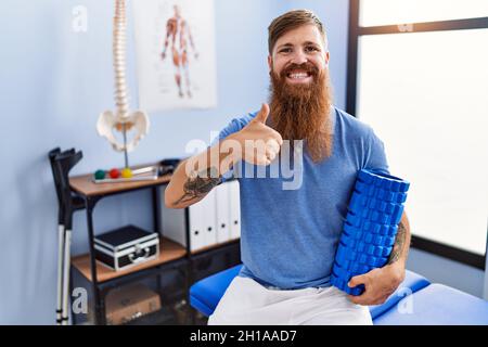 REDHEAD homme avec la longue barbe tenant le rouleau en mousse à la clinique médicale souriant heureux et positif, pouce vers le haut faisant excellent et signe d'approbation Banque D'Images