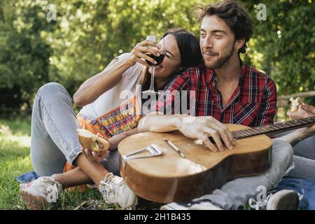 Couple de jeunes amis assis sur l'herbe en train de boire du vin et de jouer de la guitare au pique-nique Banque D'Images