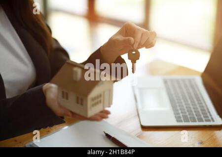 Image rognée d'une femme courtier immobilier tenant le porte-clés de la maison et le modèle de maison dans sa main, s'assoit au bureau dans le bureau. Banque D'Images