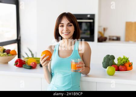 Jeune femme buvant du jus et tenant de l'orange, debout près de la table de cuisine pleine de fruits et souriant à la caméra Banque D'Images