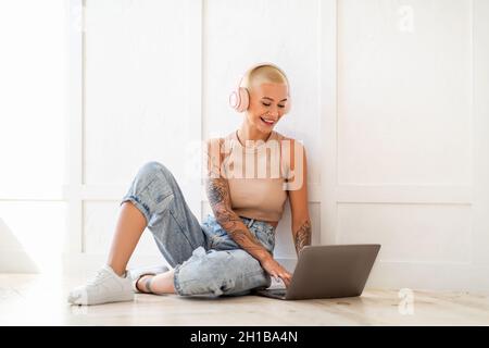 Jeune femme freelance dans les écouteurs travaillant sur ordinateur portable, assis sur le sol et s'appuyant sur le mur blanc Banque D'Images