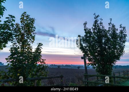 Vue grand angle du ciel de l'aube sur un paysage brumeux du Kent.Le premier plan est une porte ouverte dans une clôture en bois avec un arbre de chaque côté et un panneau « public Footpath ».Ciel bleu avec couche de rouge et de nuage mauve à l'horizon. Banque D'Images