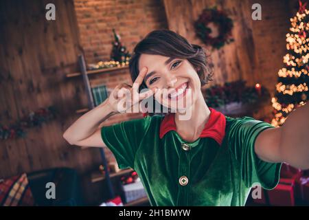 Photo d'impressionnée jolie jeune femme vêtue costume vert souriant célébrant la nouvelle année montrant V-sign intérieur chambre maison Banque D'Images