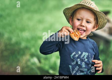 Un adorable tout-petit garçon en chapeau de paille mange une aile de poulet grillée dans une cour de campagne verte le jour de l'été, près de l'espace pour le design Banque D'Images