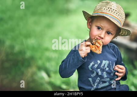 Un adorable tout-petit garçon en chapeau de paille mange une aile de poulet grillée dans une cour de campagne verte le jour de l'été, près de l'espace pour le design Banque D'Images