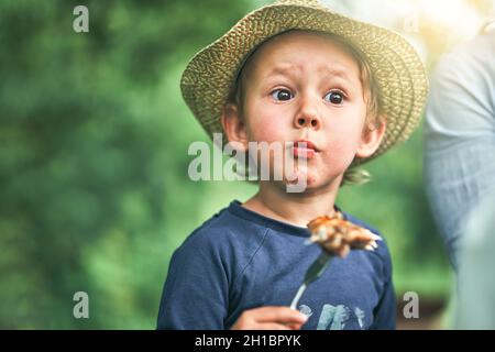 Drôle surprise petit garçon en chapeau de paille mange de délicieux ailes de poulet grillé dans le parc vert le beau jour d'été vue de près Banque D'Images