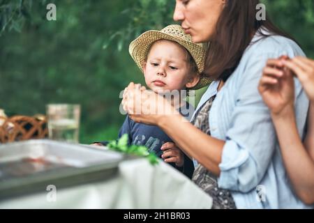 La jeune mère et le petit fils en chapeau de paille avec nourriture s'assoient à table pendant le pique-nique dans le parc vert le beau jour d'été vue de près Banque D'Images