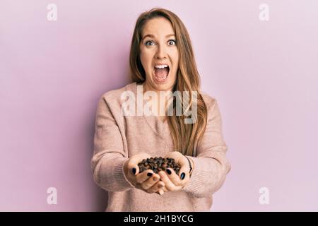 Jeune femme blonde tenant des grains de café avec les mains célébrant fou et stupéfait pour le succès avec les yeux ouverts hurlant excitée. Banque D'Images