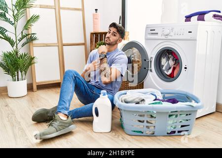 Jeune hispanique mettant sale lessive dans la machine à laver dans la hâte pointant à regarder le temps, impatience, regardant la caméra avec l'expression détendue Banque D'Images