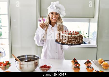 Jeune chef de pâtisserie caucasienne femme cuisant des pâtisseries et des gâteaux à la cuisine surpris par une idée ou une question pointant du doigt avec le visage heureux, numéro Banque D'Images