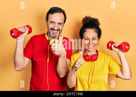 Couple d'âge moyen de femme hispanique et d'homme portant des vêtements de sport en utilisant des haltères souriant heureux et positif, pouce vers le haut faisant excellent et signe d'approbation Banque D'Images