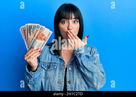 Une jeune femme hispanique détient des billets en livres égyptiennes couvrant la bouche avec la main, choquée et effrayée par erreur. Expression surprise Banque D'Images