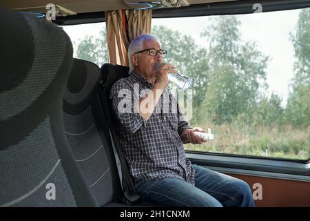 Homme senior prenant la pilule et la boire par l'eau tout en étant assis par la fenêtre sur le siège double dans le bus Banque D'Images