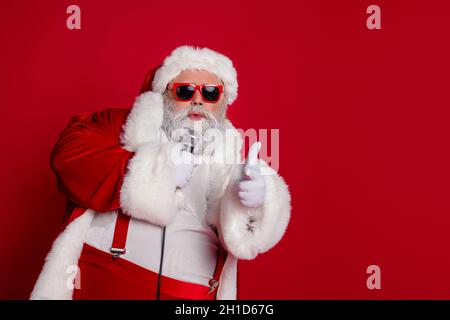 Photo de cool drôle santa claus porter rouge costume sombre lunettes chantant karaoké pointant le doigt vous vide espace isolé rouge couleur arrière-plan Banque D'Images