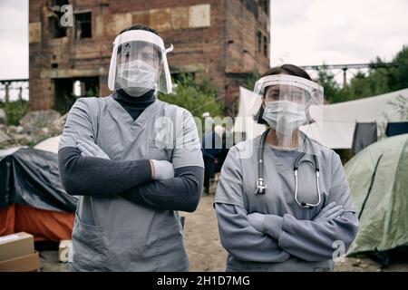 Deux jeunes clincians en vêtements de travail de protection se tenant contre des tentes de migrants dans le camp de réfugiés Banque D'Images