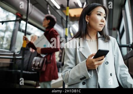 Portrait d'une élégante femme asiatique regardant la fenêtre dans le bus tout en voyageant en transports en commun et en tenant un smartphone, un espace de copie Banque D'Images