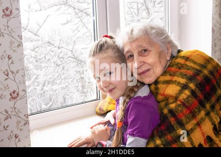 Bonne belle grand-mère et sa adorable petite-fille assis à la maison avec une grande fenêtre en hiver Banque D'Images